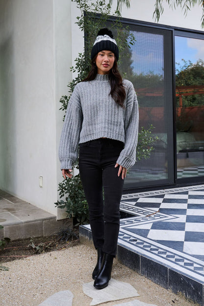 Woman in a Grey Wide Sleeve Ribbed Mock Neck Sweater, black jeans, boots, and beanie stands by a modern building with large windows.