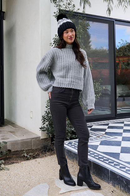 A woman wears a Grey Wide Sleeve Ribbed Mock Neck Sweater with black jeans and boots, standing outside by a modern patio.