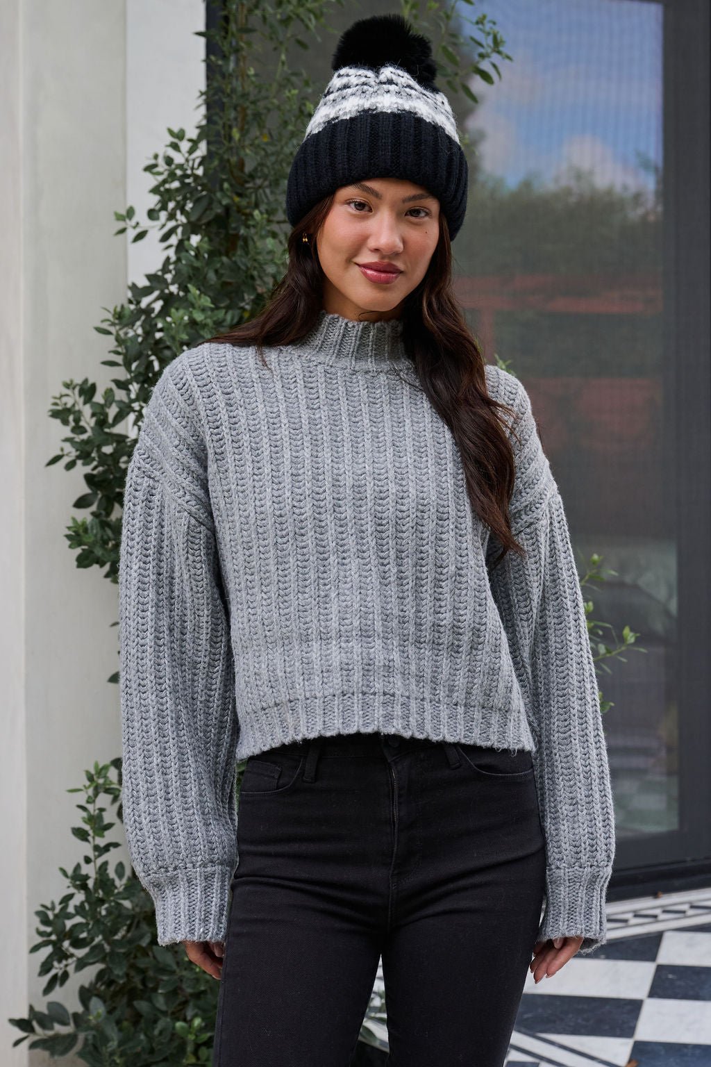 A woman wearing a Grey Wide Sleeve Ribbed Mock Neck Sweater, black pants, and a black winter hat stands outside, smiling.