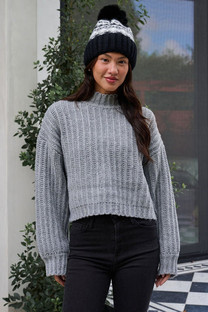 A woman wearing a Grey Wide Sleeve Ribbed Mock Neck Sweater, black pants, and a black winter hat stands outside, smiling.