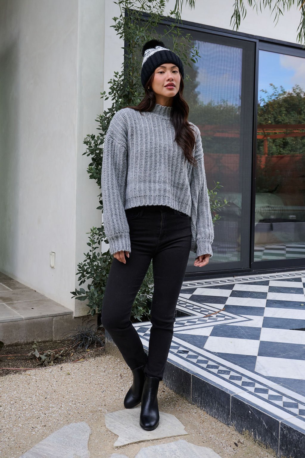 A woman in a Grey Wide Sleeve Ribbed Mock Neck Sweater, black pants, and a beanie stands outside on a tiled patio by glass doors.