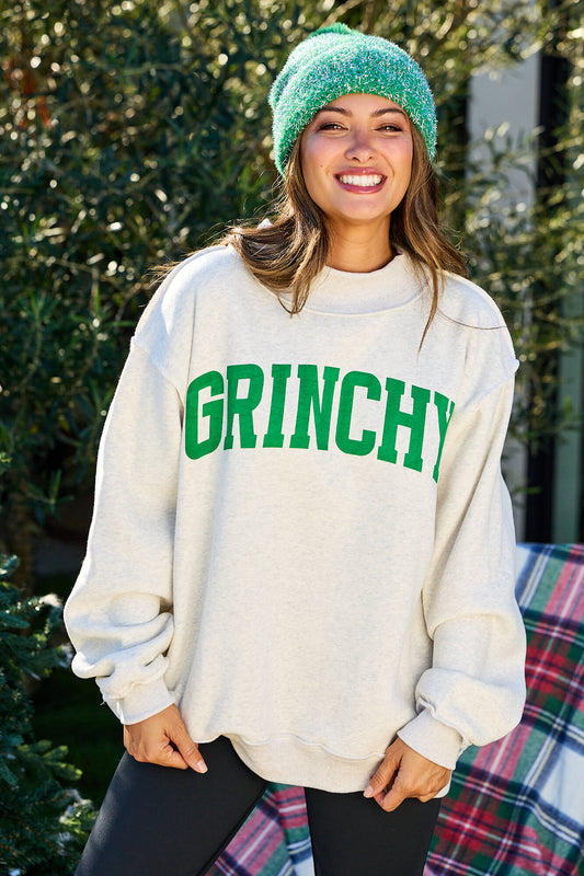 Smiling woman in a Grinchy/Naughty Reversible Oversized Sweatshirt and green beanie stands outside—festive holiday outfit vibes.