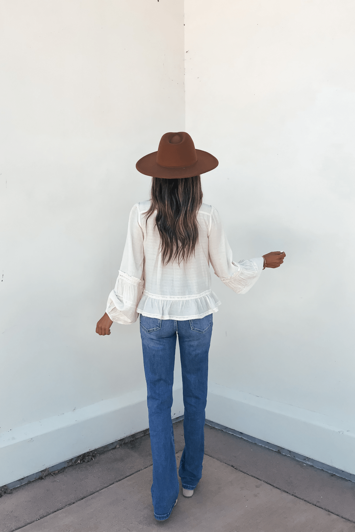 Someone in a brown hat and blue jeans faces a white wall, wearing the Hadley Cream Ruffle Lace Trim Blouse, photographed from behind.