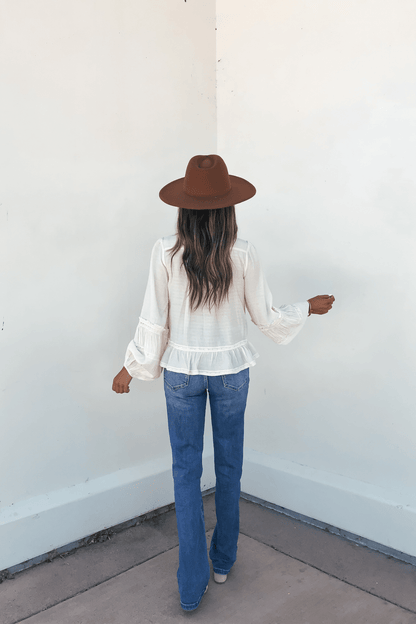 Someone in a brown hat and blue jeans faces a white wall, wearing the Hadley Cream Ruffle Lace Trim Blouse, photographed from behind.