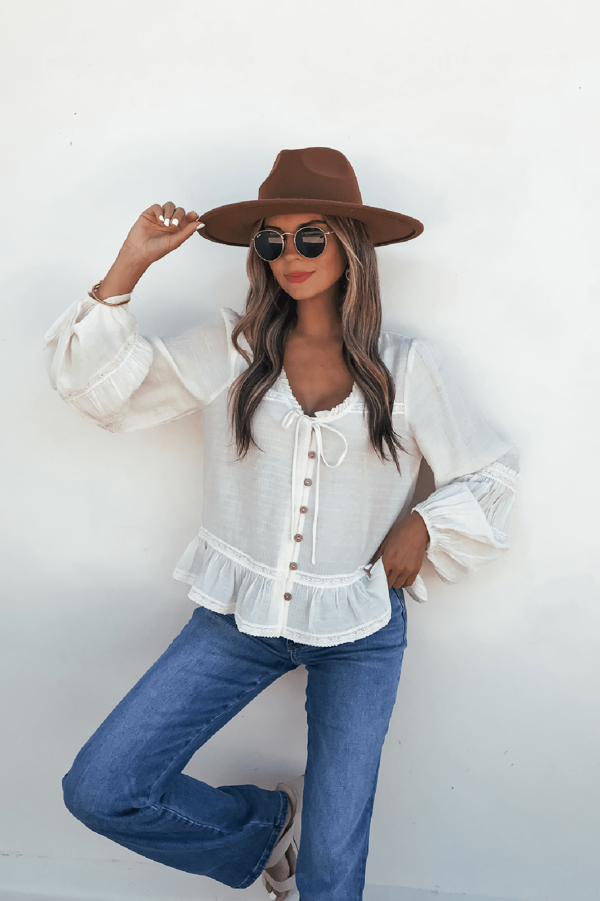 Woman in a brown hat, Hadley Cream Ruffle Lace Trim Blouse, blue jeans, sunglasses, and sandals poses against a white wall.