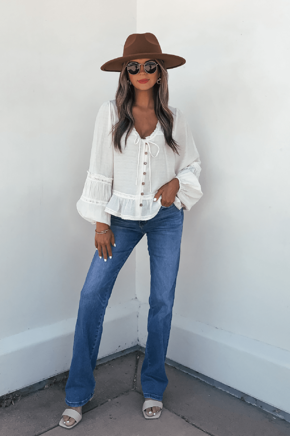 Woman in hat, sunglasses, Hadley Cream Ruffle Lace Trim Blouse, blue jeans, and sandals poses by a white corner wall.