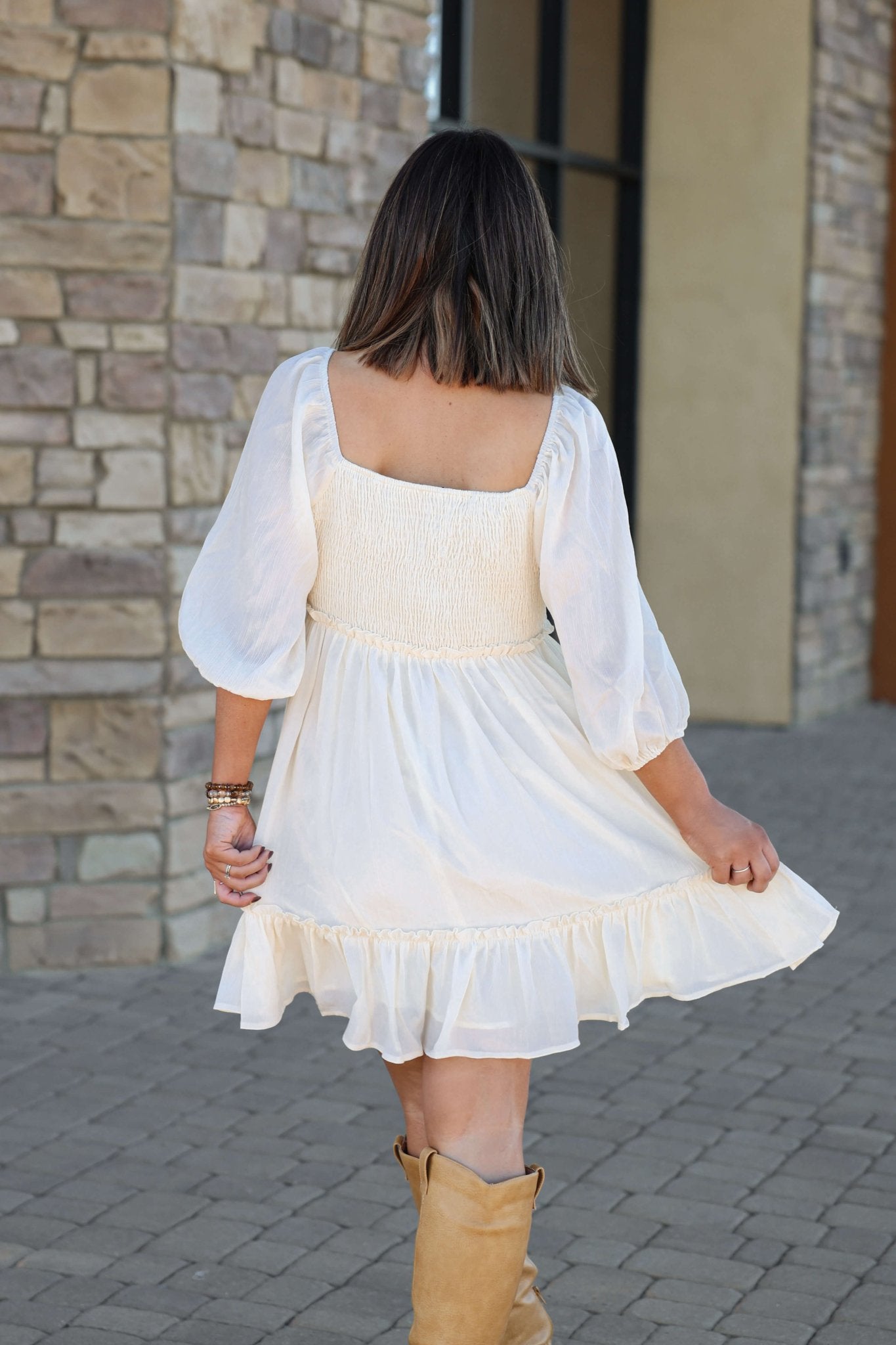 Woman in a bump-friendly Hadley Cream Smocked Mini Dress - DOORBUSTER with tan boots walks on a stone path near a stone wall.