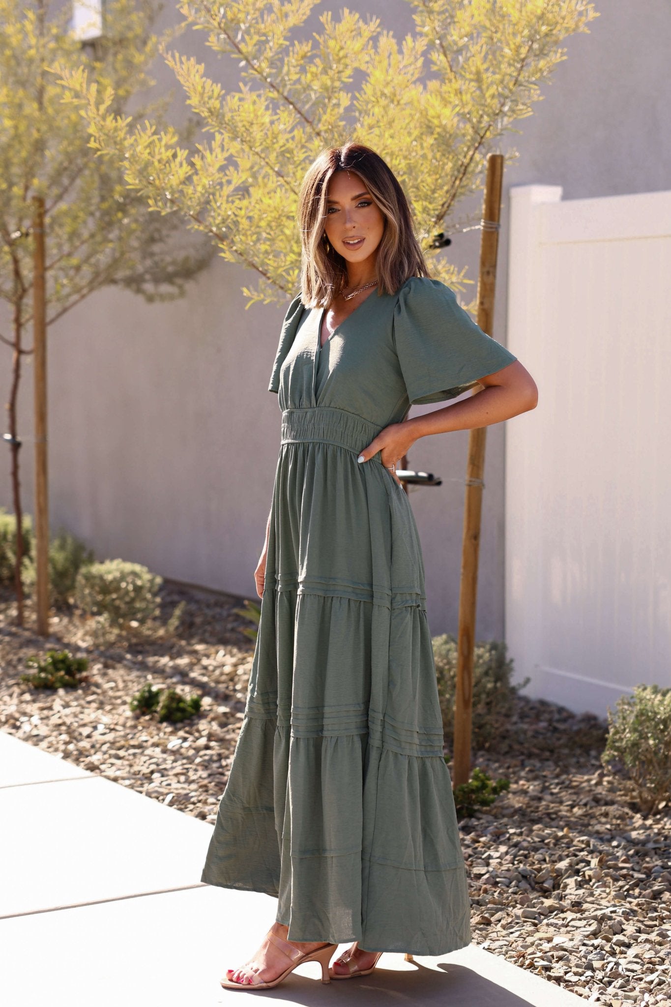 Woman in an olive Hannah Surplice Tiered Maxi Dress stands outdoors on a sunny day, with trees and shrubs in the background.