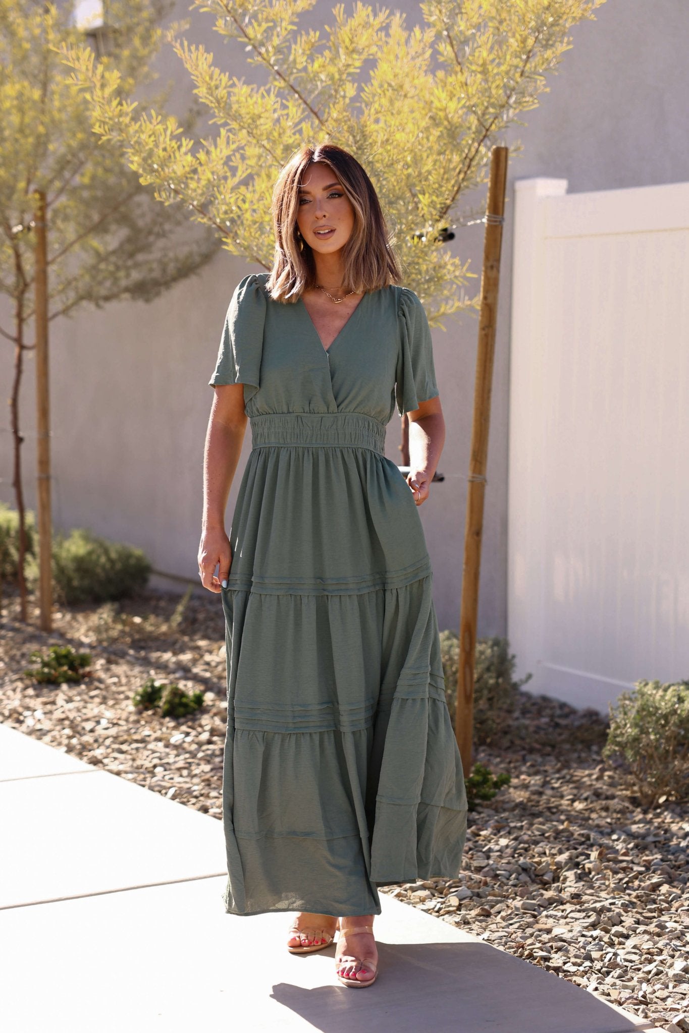 Woman in the Hannah Surplice Tiered Maxi Dress - Olive, tan sandals, and light brown hair stands outside on a sunny day.