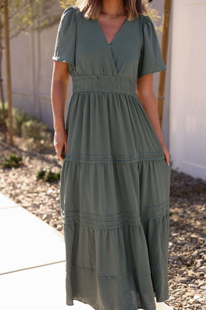 A woman wears the Hannah Surplice Tiered Maxi Dress in Olive outdoors on a sunny day.