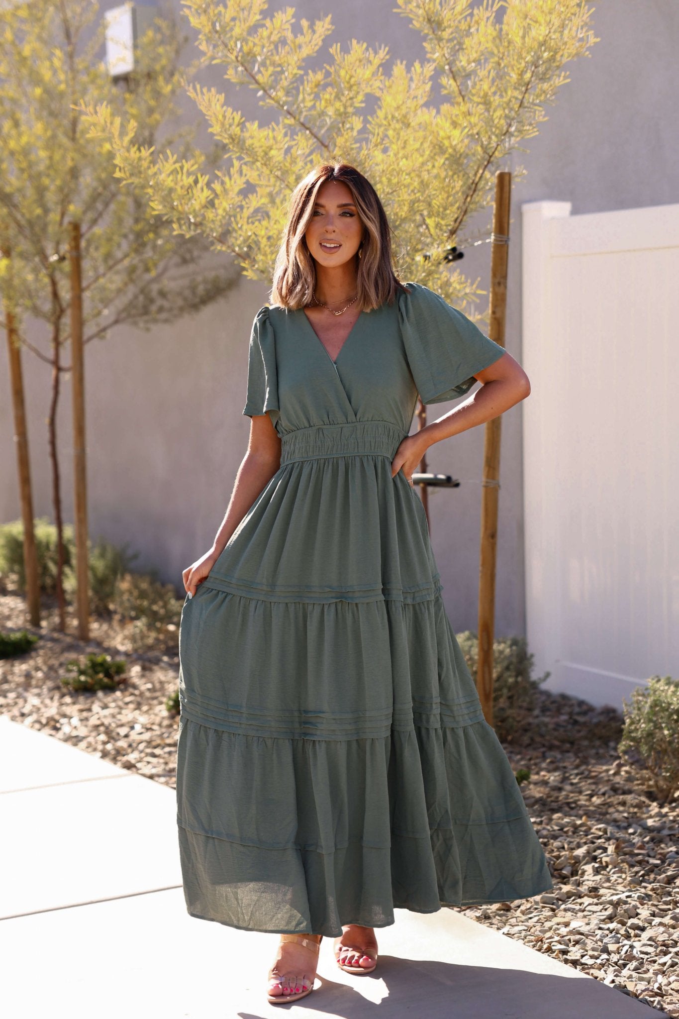 Woman smiles outdoors in the Hannah Surplice Tiered Maxi Dress - Olive, holding her flowing dress in the sunlight.