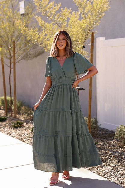 Woman smiles outdoors in the Hannah Surplice Tiered Maxi Dress - Olive, holding her flowing dress in the sunlight.