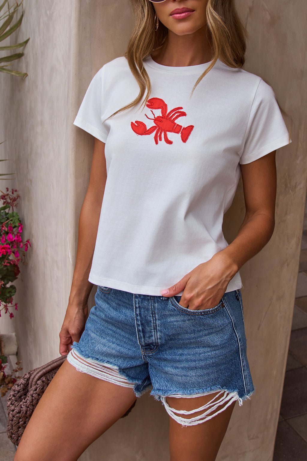 Woman in the Harbor Lobster Patch Tee and distressed shorts stands by a beige wall, creating a coastal vibe.