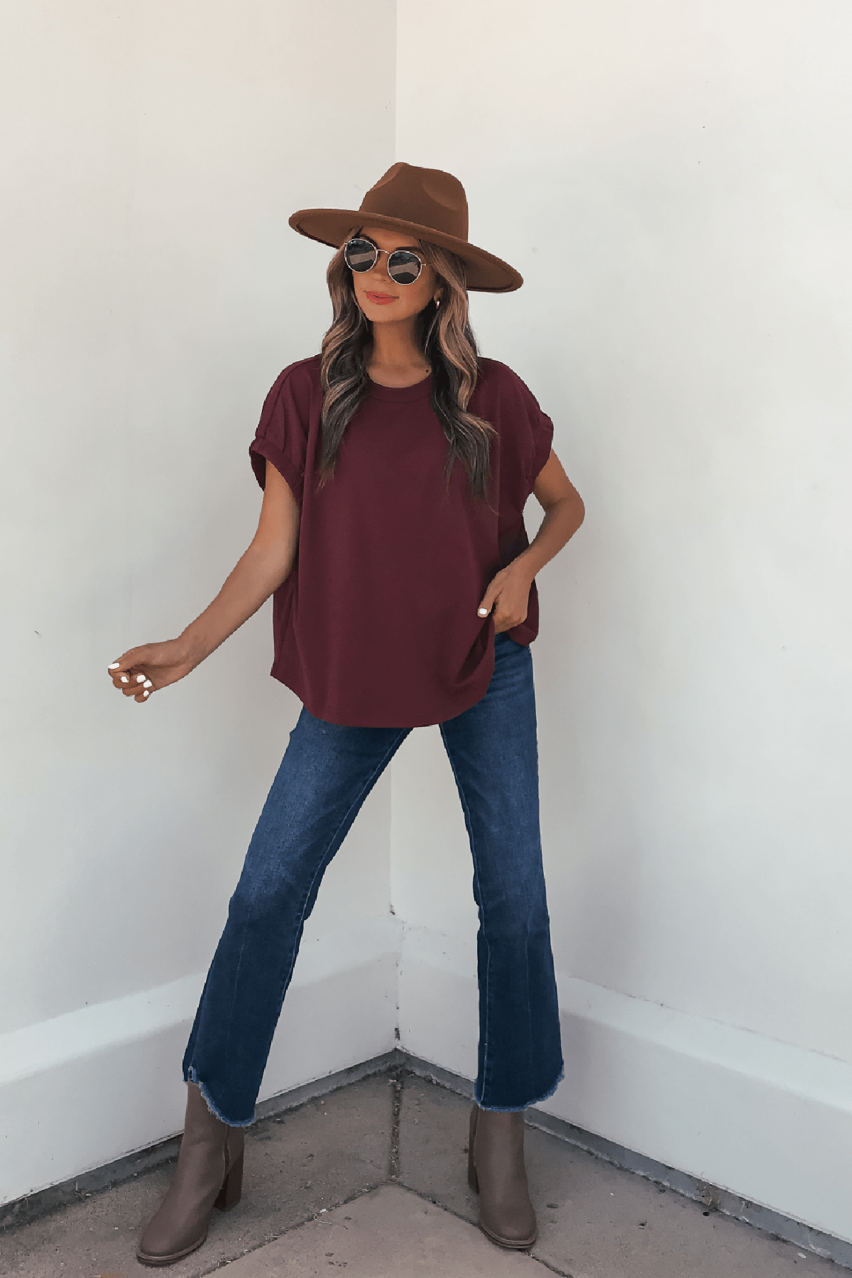 Woman in sunglasses, Harlow Burgundy Short Sleeve Blouse, blue jeans, brown hat and boots stands against a white wall.
