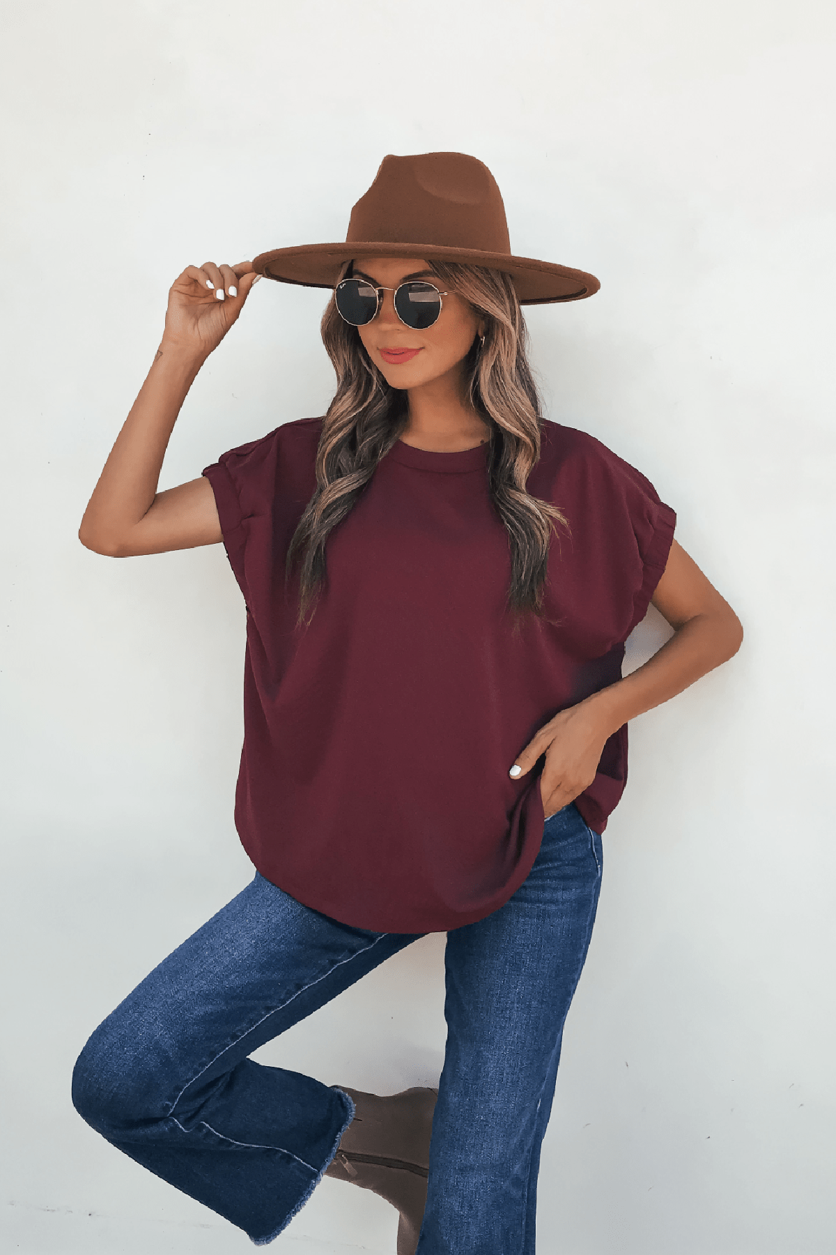 Woman wearing a brown hat, sunglasses, Harlow Burgundy Short Sleeve Blouse, and blue jeans poses against a white background.