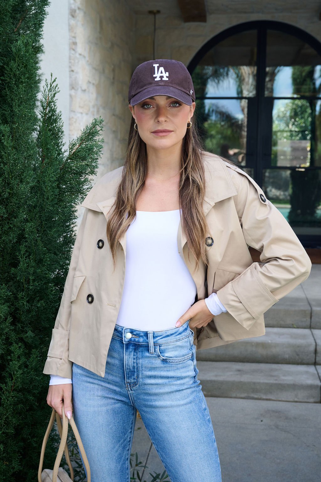 A woman outdoors by a stone building wears the Harper Beige Trench Coat, white top, blue jeans, LA cap, and holds a beige bag.