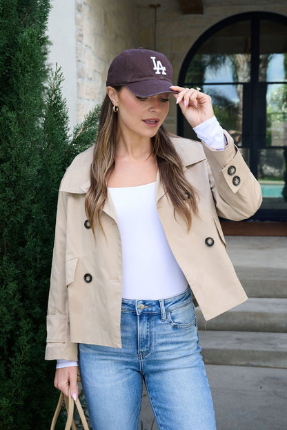 Woman in a Harper Beige Trench Coat, white top, and blue jeans adjusts a brown LA cap outdoors with a beige bag in hand.