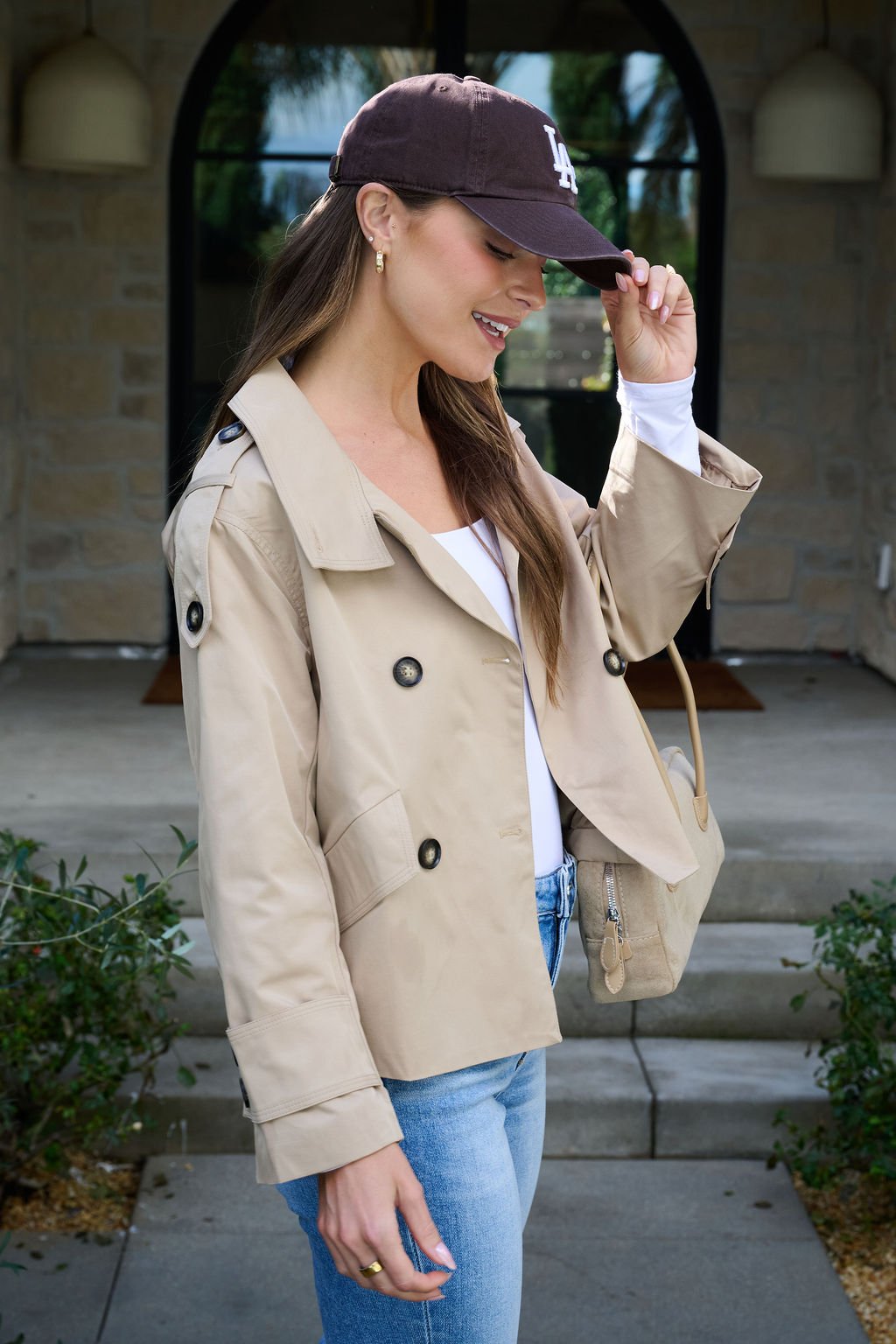 A woman in a Harper Beige Trench Coat, white top, and jeans smiles while adjusting her cap outside a stone building.