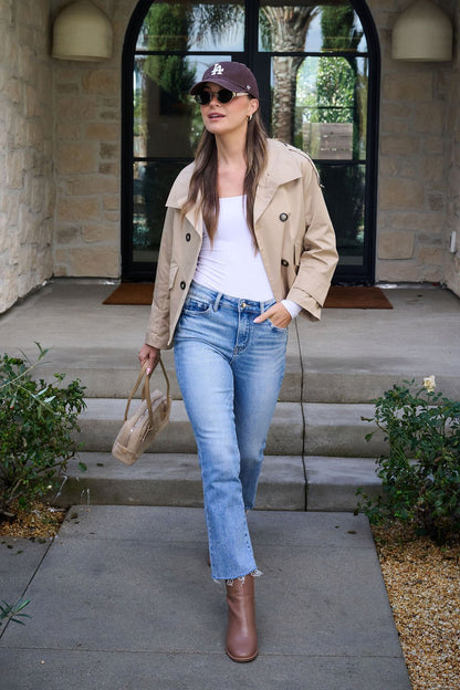 A woman in jeans and a white top walks outside in the Harper Beige Trench Coat, paired with brown boots and a beige handbag.