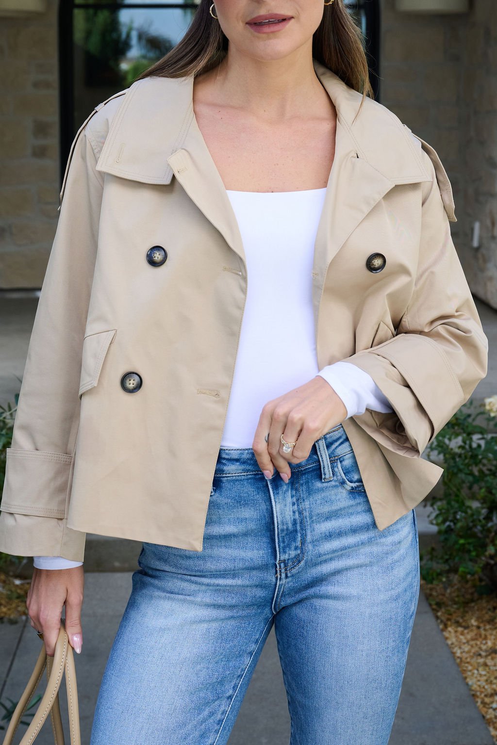 Woman in the Harper Beige Trench Coat with a white top, blue jeans, and tan bag, standing outside by a stone building.