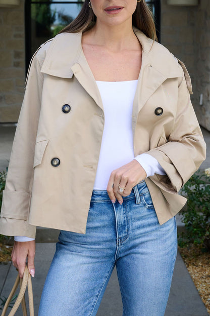 Woman in the Harper Beige Trench Coat with a white top, blue jeans, and tan bag, standing outside by a stone building.