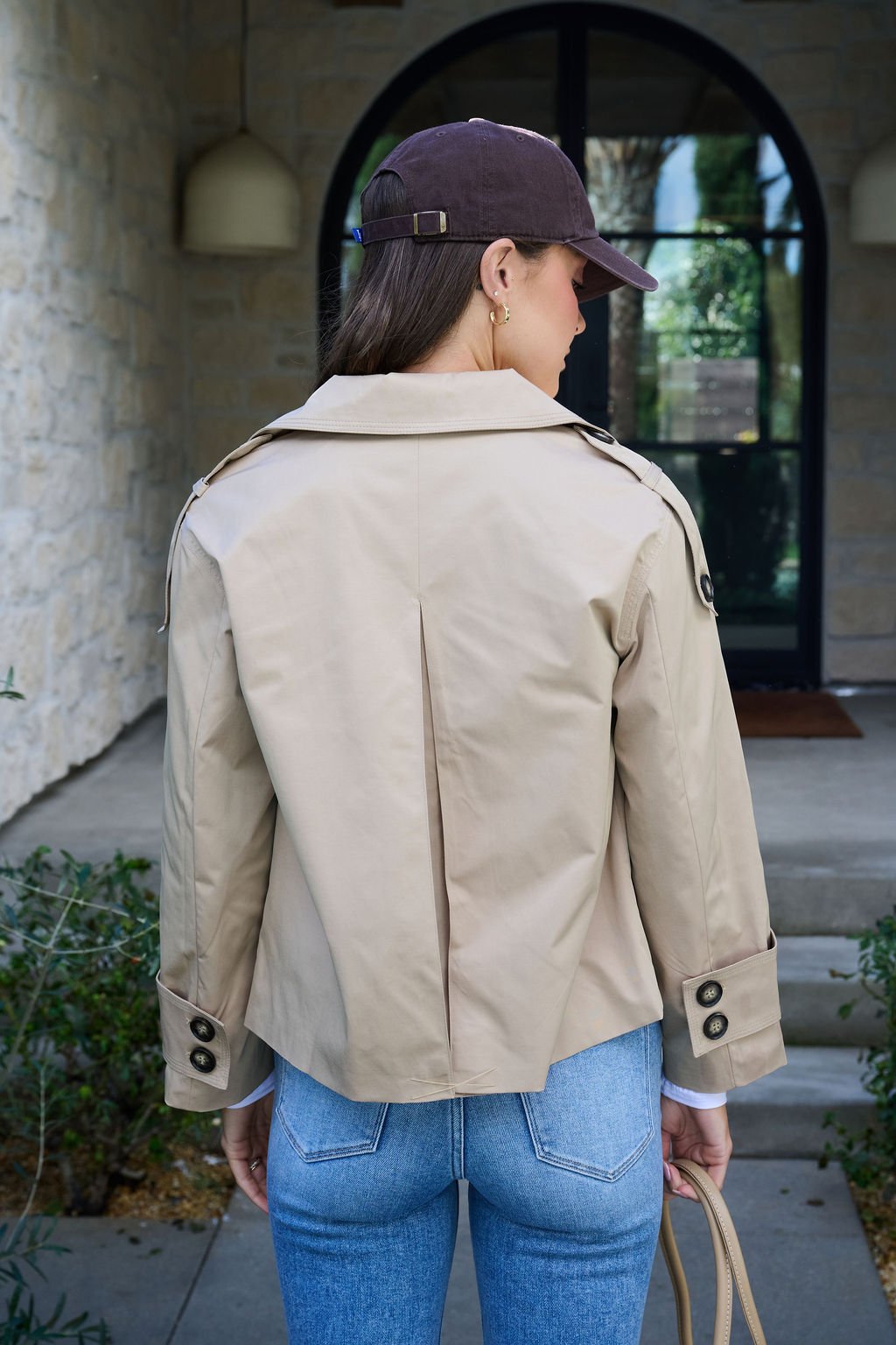 A woman in a Harper Beige Trench Coat, blue jeans, and a dark cap stands outside a building with her back to the camera.