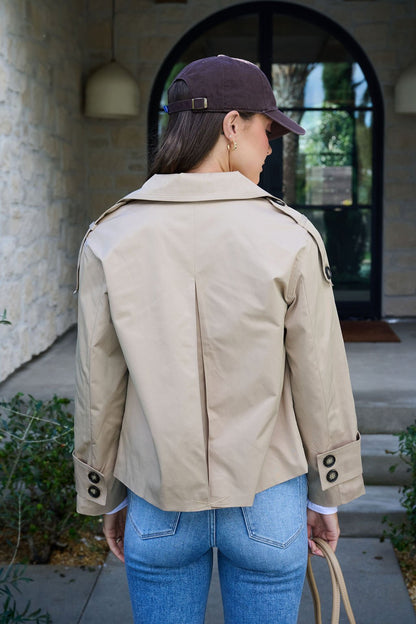 A woman in a Harper Beige Trench Coat, blue jeans, and a dark cap stands outside a building with her back to the camera.