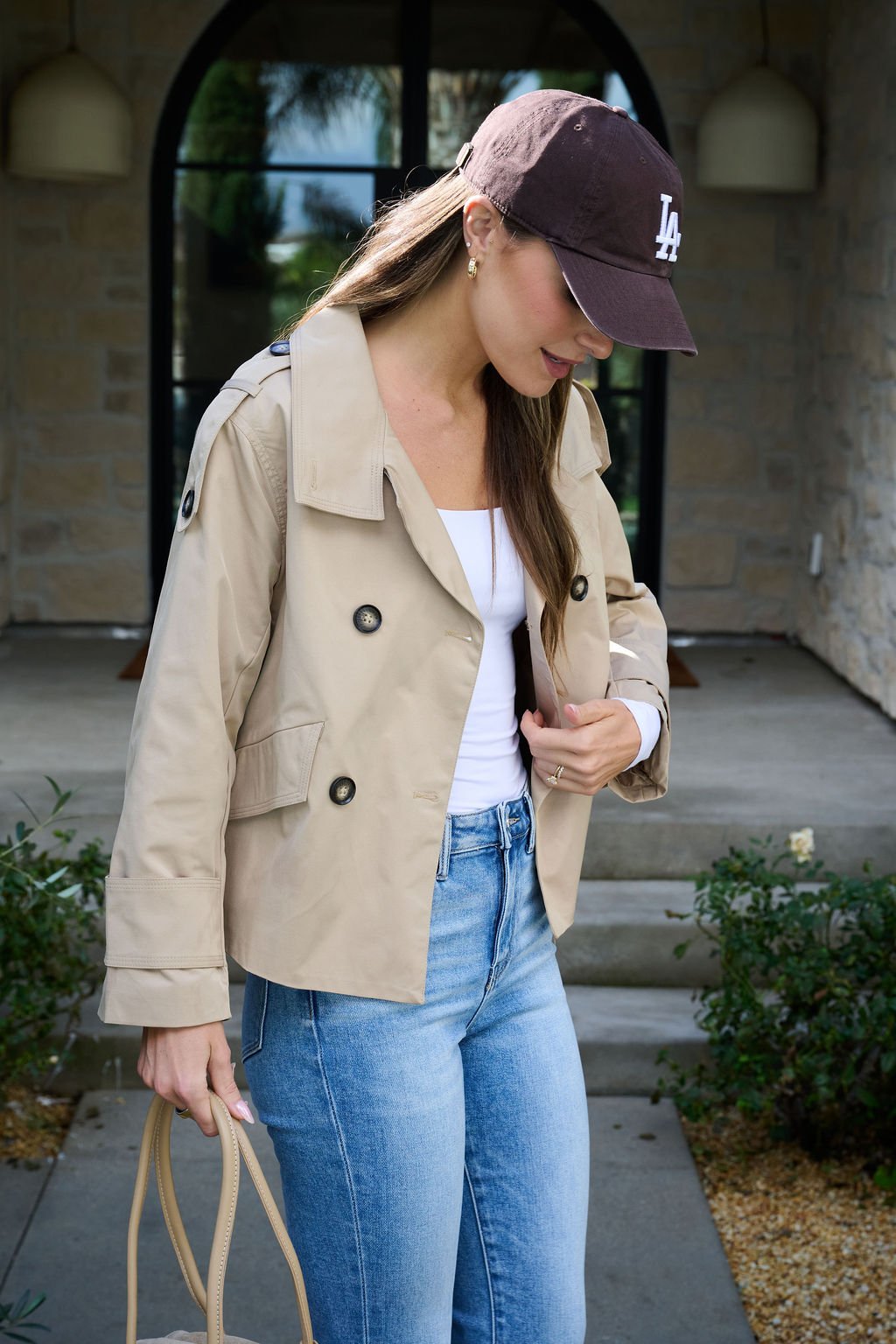 Wearing a Harper Beige Trench Coat, blue jeans, and an LA cap, she stands outside holding a beige bag and looks down.