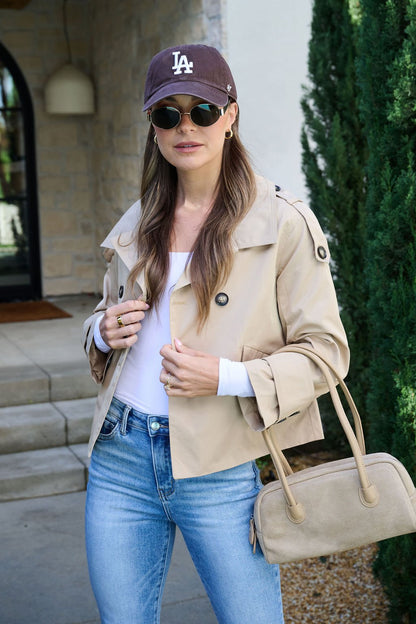 A woman in a Harper Beige Trench Coat, blue jeans, and LA cap stands outside with a matching bag and sunglasses.