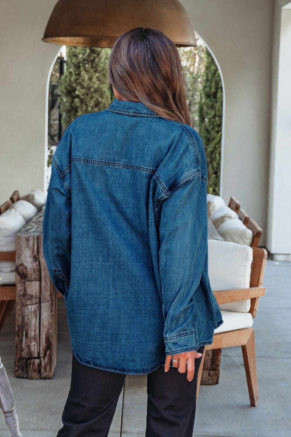 A woman wears the Harvest Blues Dark Denim Shacket–FINAL SALE, perfect for fall, standing with her back on a patio with wooden furniture.