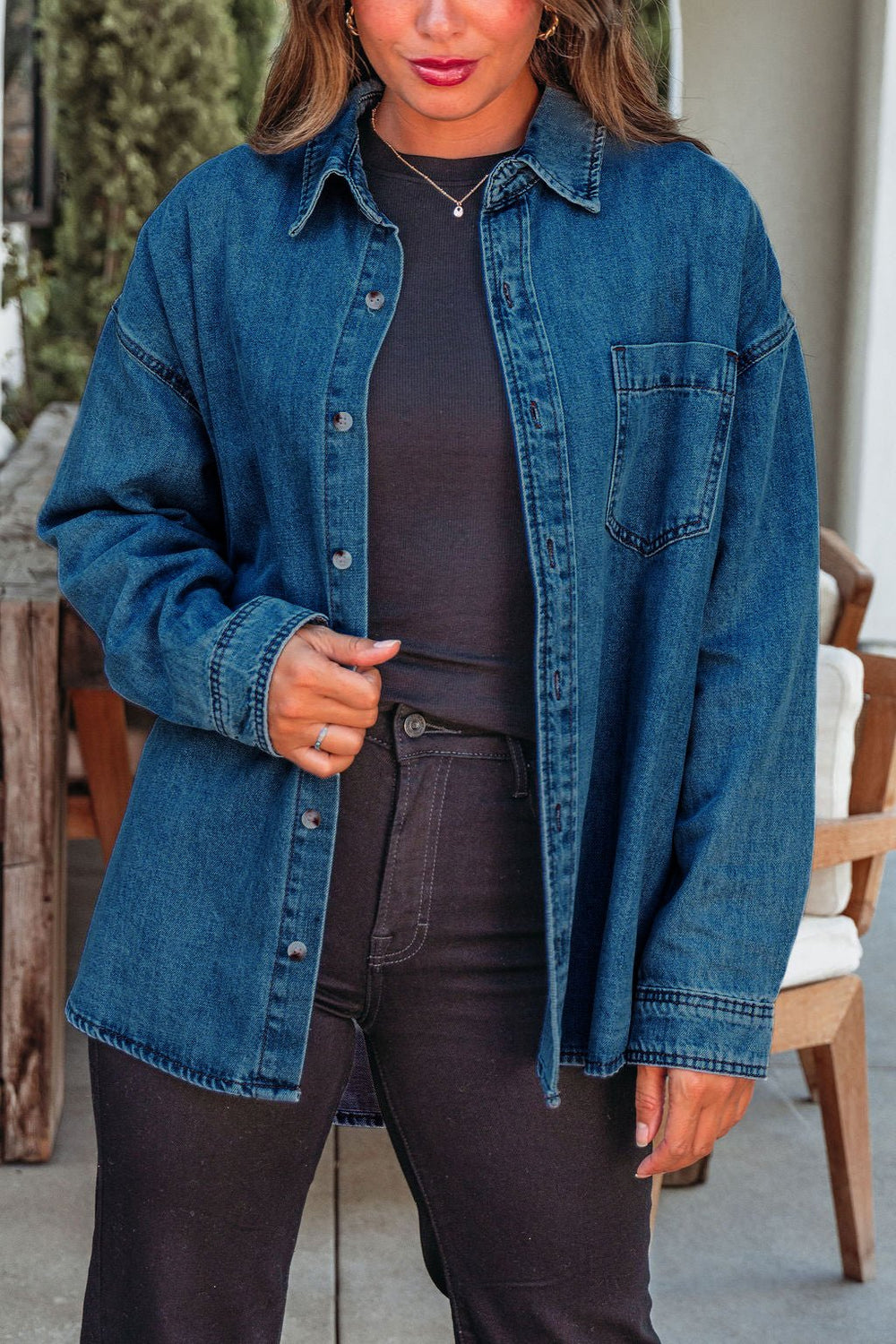 The Harvest Blues Dark Denim Shacket, 100% cotton, layers stylishly over black jeans for fall by wooden outdoor furniture.