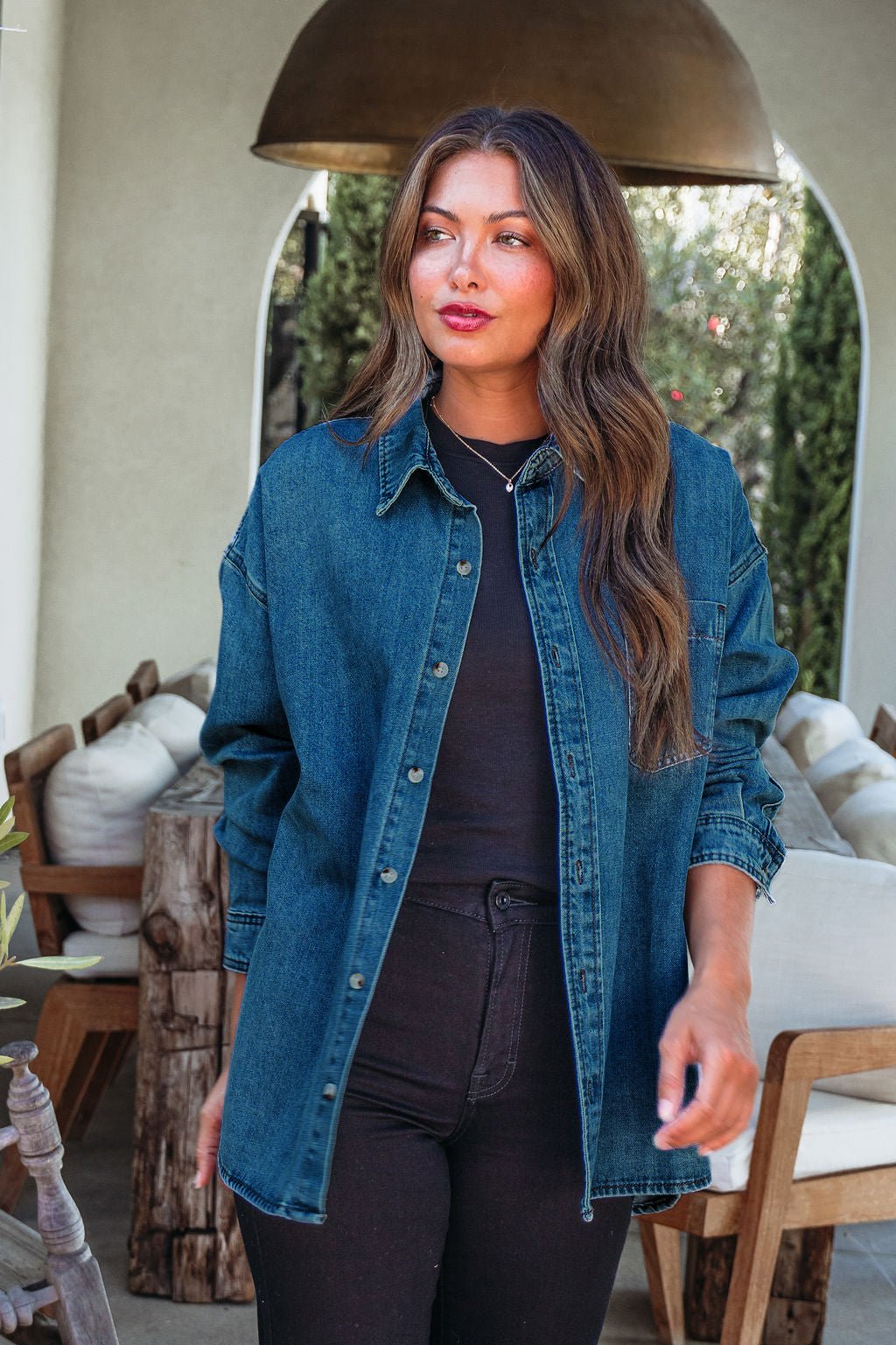 Woman with long brown hair wearing the Harvest Blues Dark Denim Shacket over black, standing in a stylish outdoor lounge.