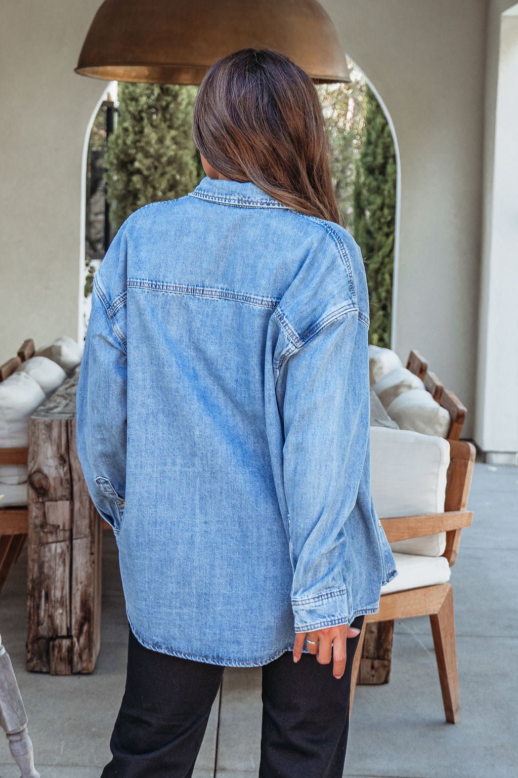 A person with long brown hair wears the Harvest Blues Denim Shacket and black pants, standing with their back to the camera on a patio.