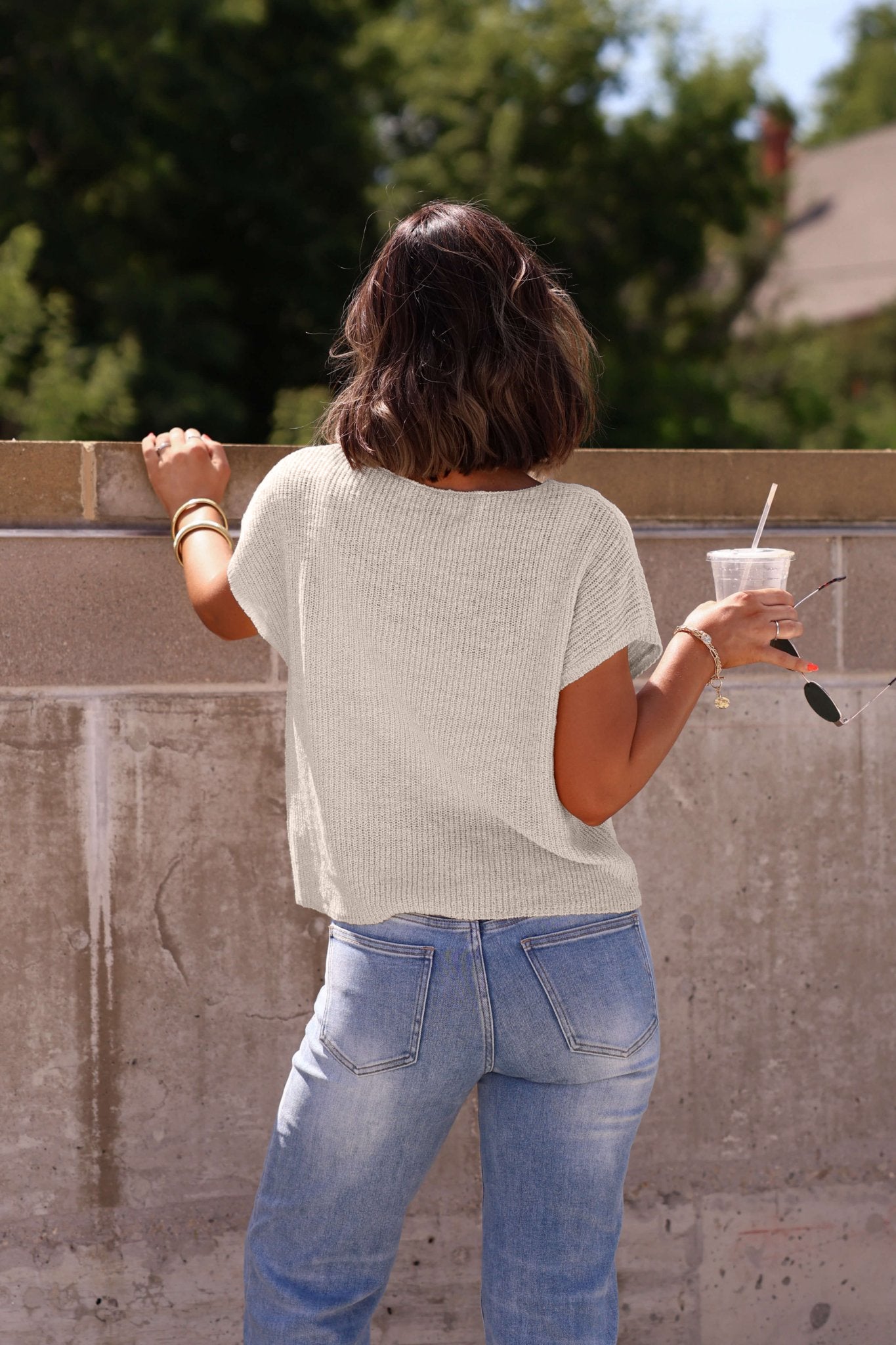 A woman in jeans and the Haven Natural Cap Sleeve Sweater stands by a concrete wall, holding a drink and sunglasses, facing away.