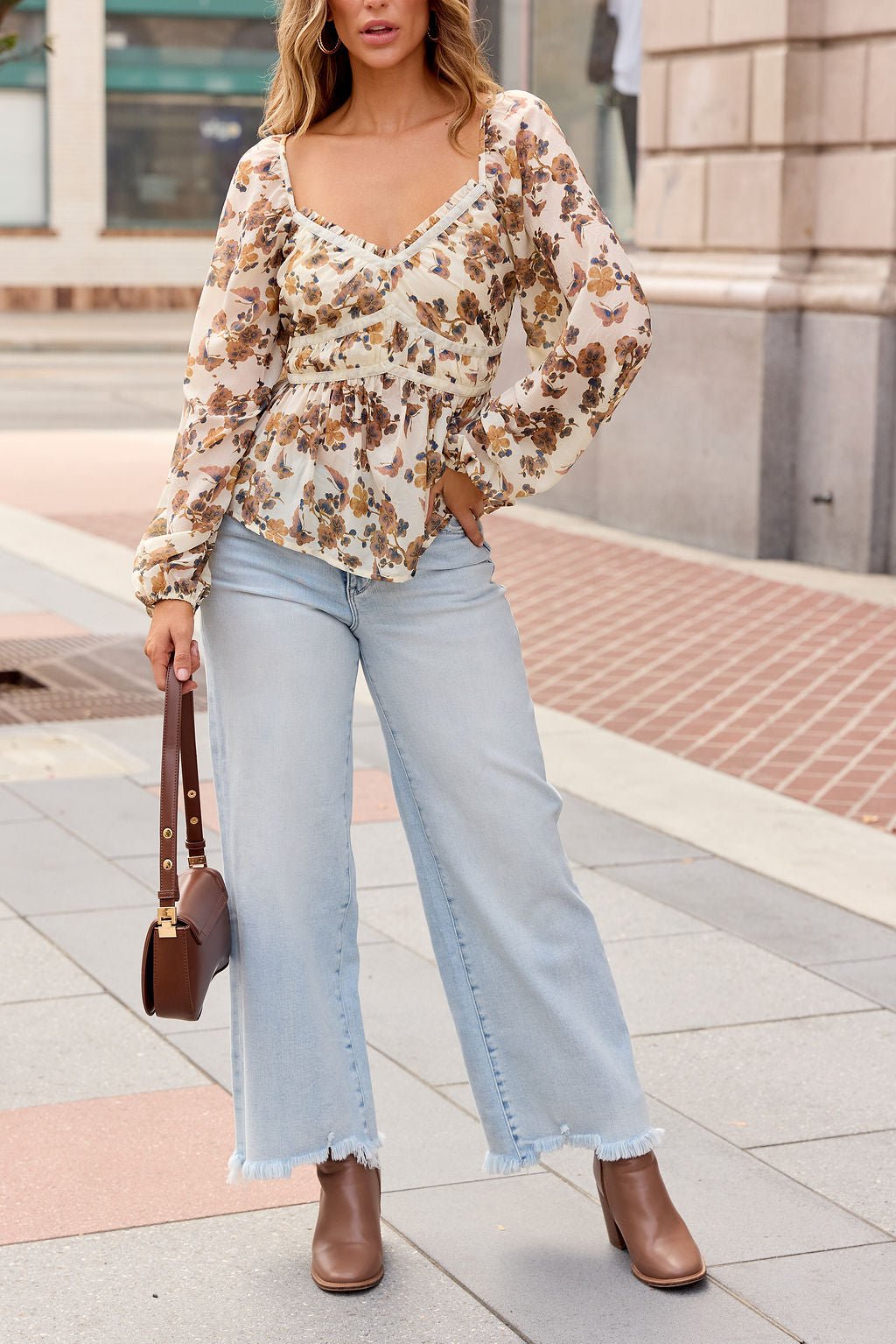 Woman in the Hazel Floral Print Top - Cream stands on a city sidewalk, styled with jeans, brown boots, and a matching handbag.