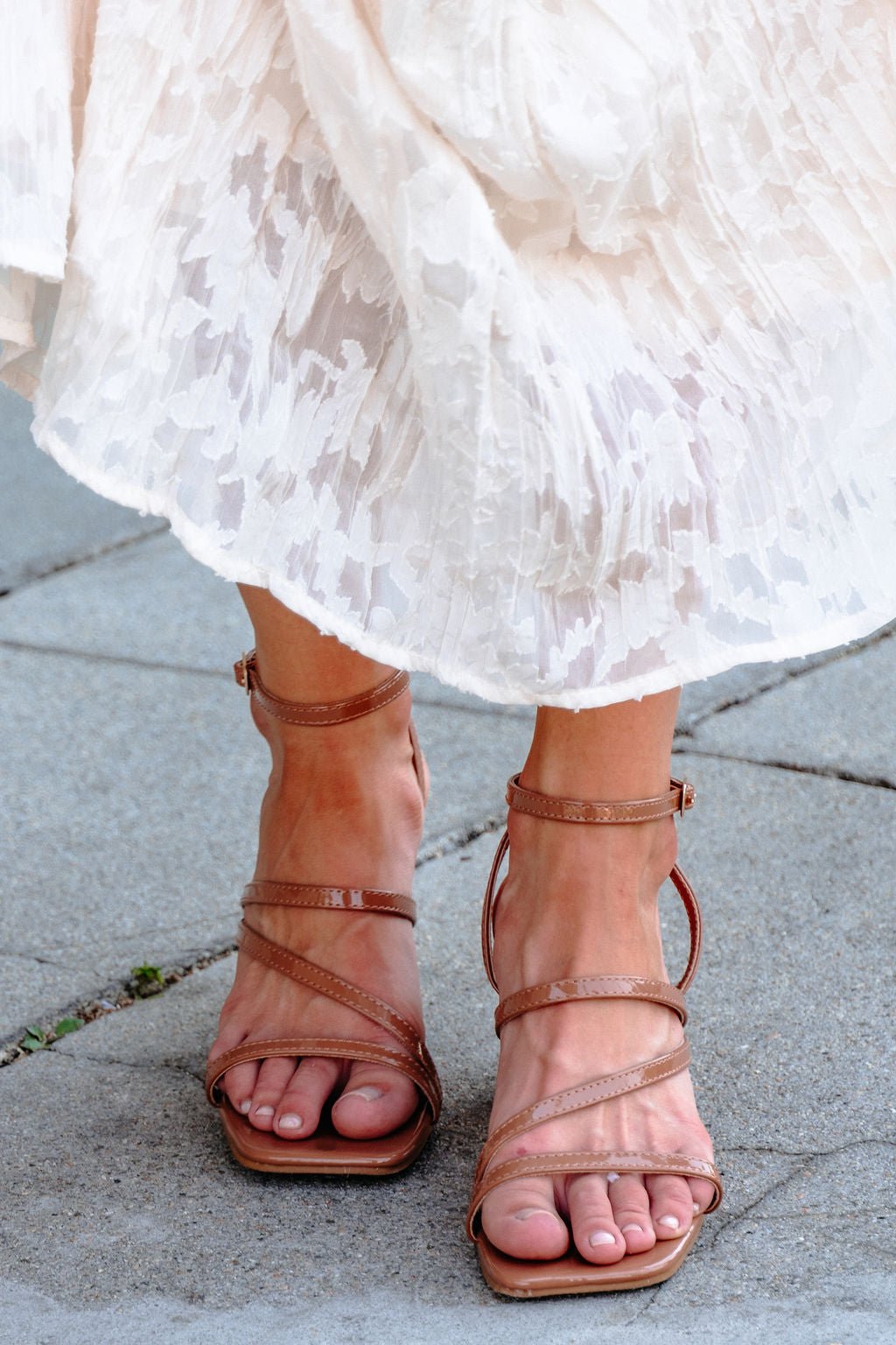 Someone wears Hazel Strappy Heeled Sandals - DOORBUSTER with a white textured skirt, standing on stone pavement.