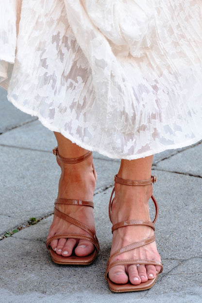 Someone wears Hazel Strappy Heeled Sandals - DOORBUSTER with a white textured skirt, standing on stone pavement.