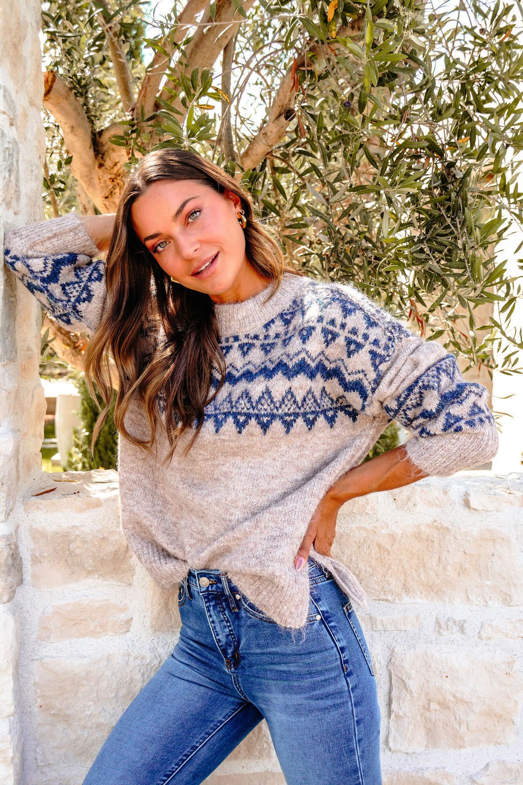 A woman in a Heather Grey and Blue Aztec Print Sweater and jeans poses by a stone wall, smiling with one arm raised.
