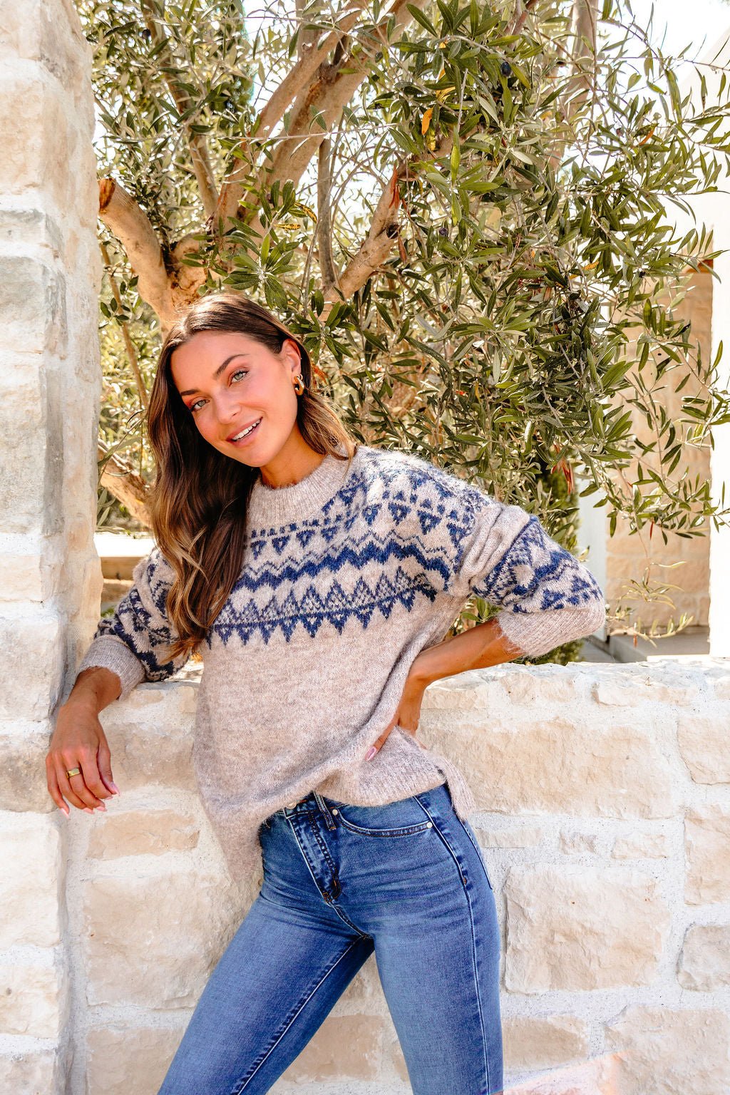 Woman wears a Heather Grey and Blue Aztec Print Sweater and jeans, leaning on a stone wall with olive trees in the background.