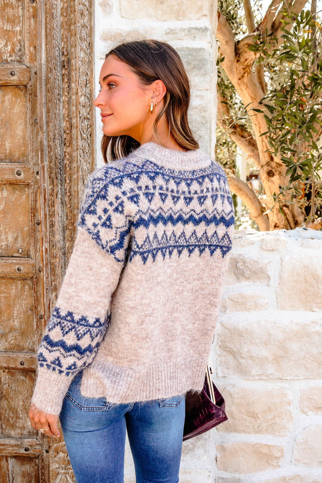 Woman in a Heather Grey and Blue Aztec Print Sweater and jeans stands outdoors by a rustic wooden door and stone wall.