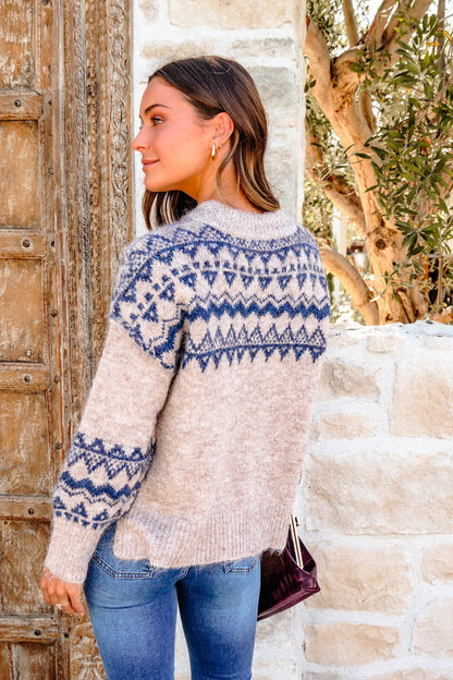 Woman in a Heather Grey and Blue Aztec Print Sweater and jeans stands outdoors by a rustic wooden door and stone wall.