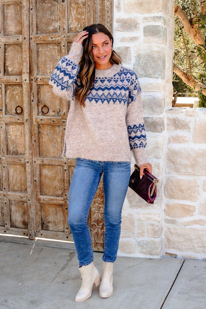 Woman in a Heather Grey and Blue Aztec Print Sweater, jeans, and boots by a rustic wooden door and stone wall.