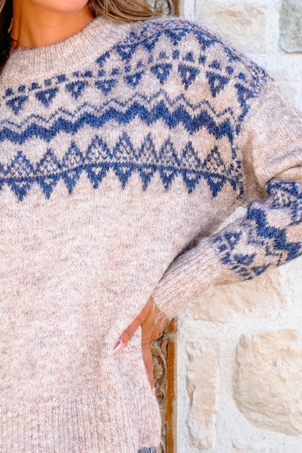 Wearing a Heather Grey and Blue Aztec Print Sweater, a person stands by a light stone wall with hand on hip.