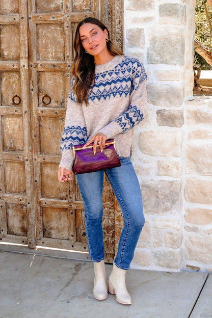 Woman in a Heather Grey and Blue Aztec Print Sweater, jeans, and white boots holds a purple clutch by a stone wall and wooden door.
