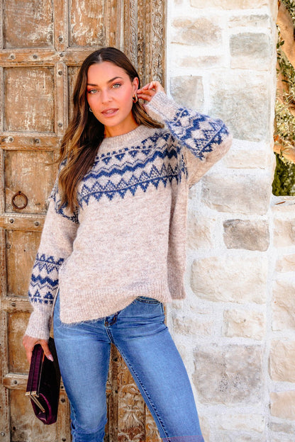 Woman in a Heather Grey and Blue Aztec Print Sweater with an oversized fit, jeans, and clutch stands by a rustic wooden door.