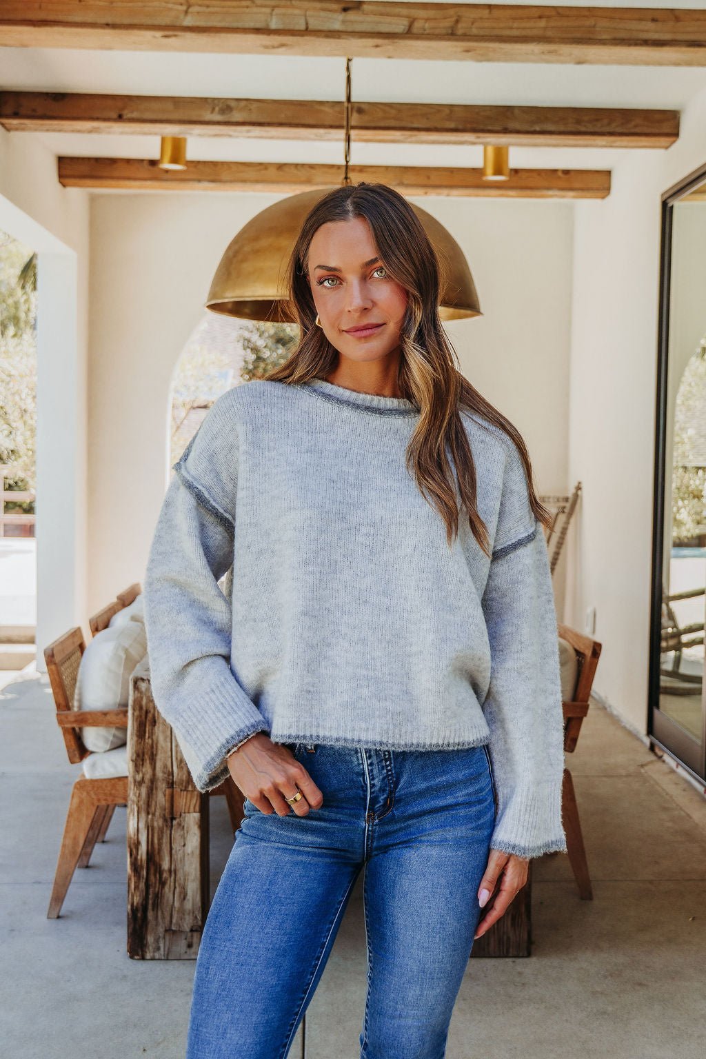 A woman wears the Heather Grey Contrast Seam Detail Sweater and blue jeans, smiling indoors with rustic dining décor and warm lighting.