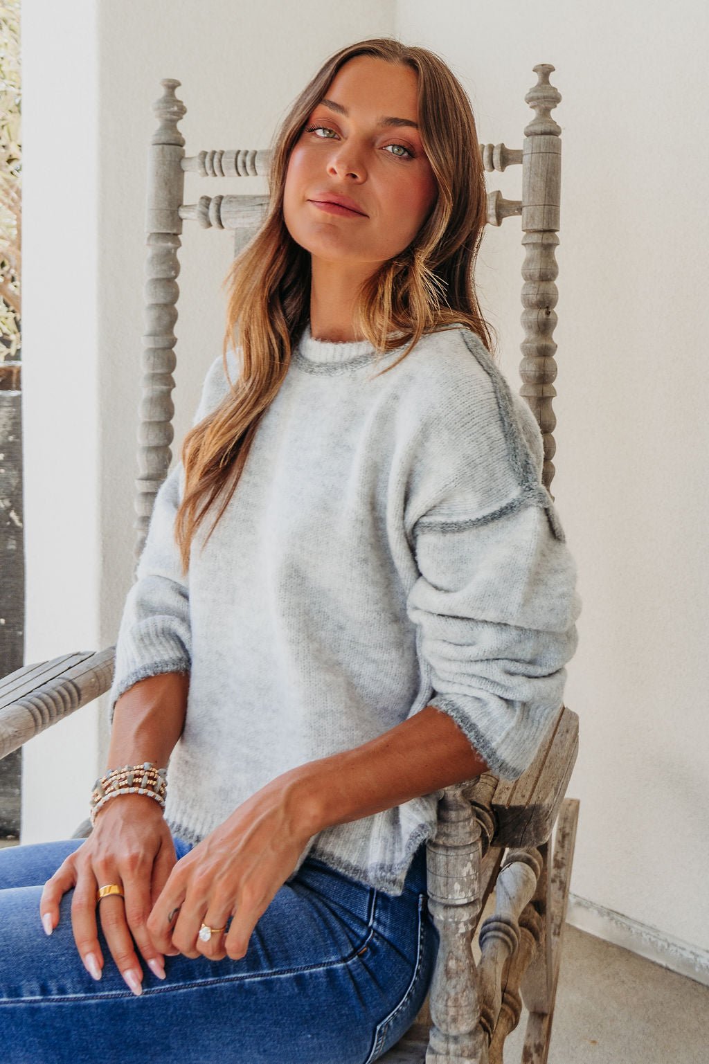 Woman with long brown hair wears a Heather Grey Contrast Seam Detail Sweater and jeans, sitting on a wooden chair in bright indoor light.