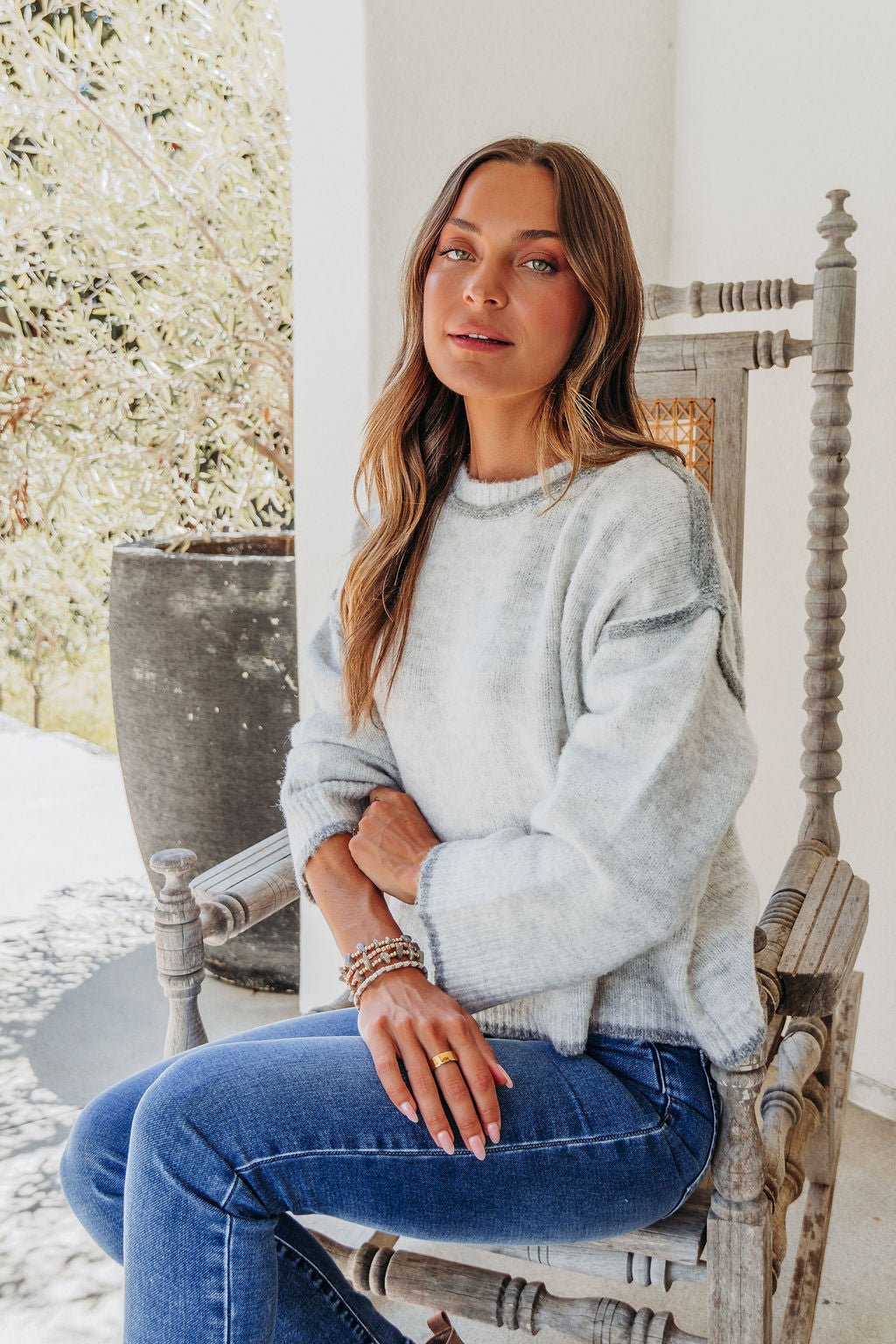 A woman with long hair sits on a wooden chair, wearing the Heather Grey Contrast Seam Detail Sweater and blue jeans.
