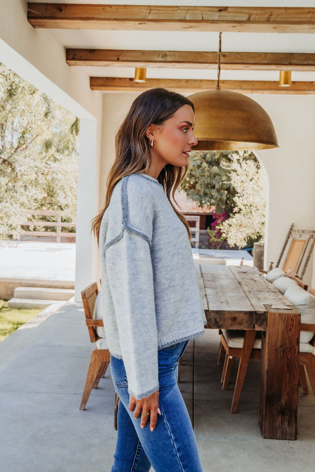 A woman in a Heather Grey Contrast Seam Detail Sweater and blue jeans stands in profile on a patio next to a wooden dining set.