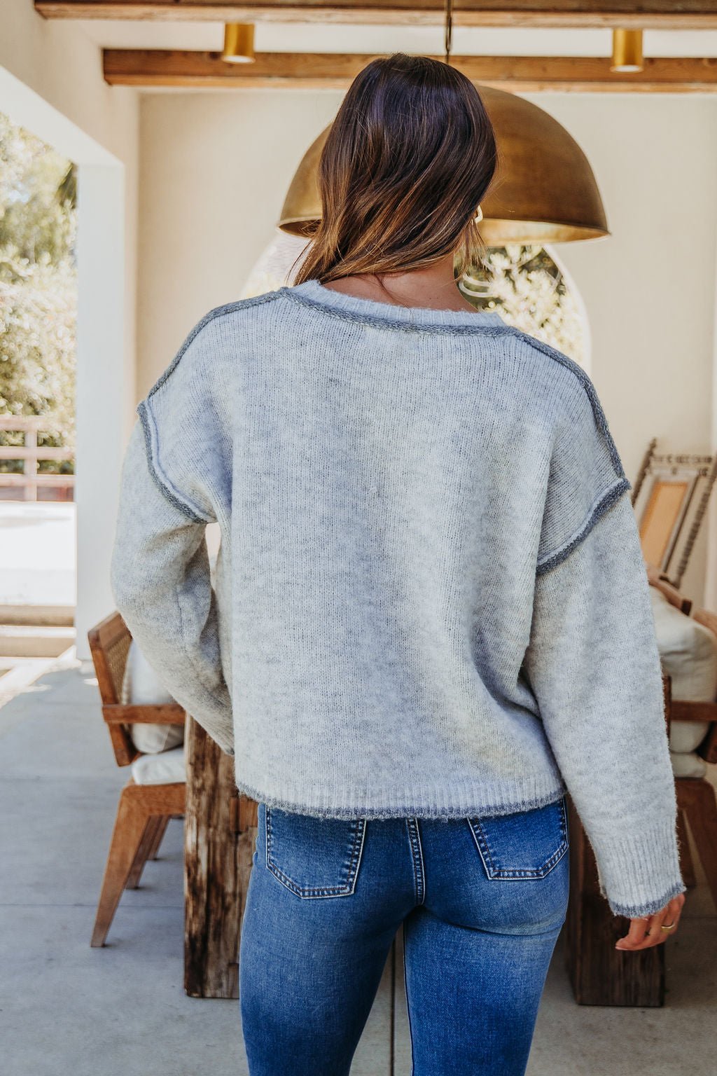 Woman standing indoors with her back to the camera, wearing a Heather Grey Contrast Seam Detail Sweater and blue jeans.
